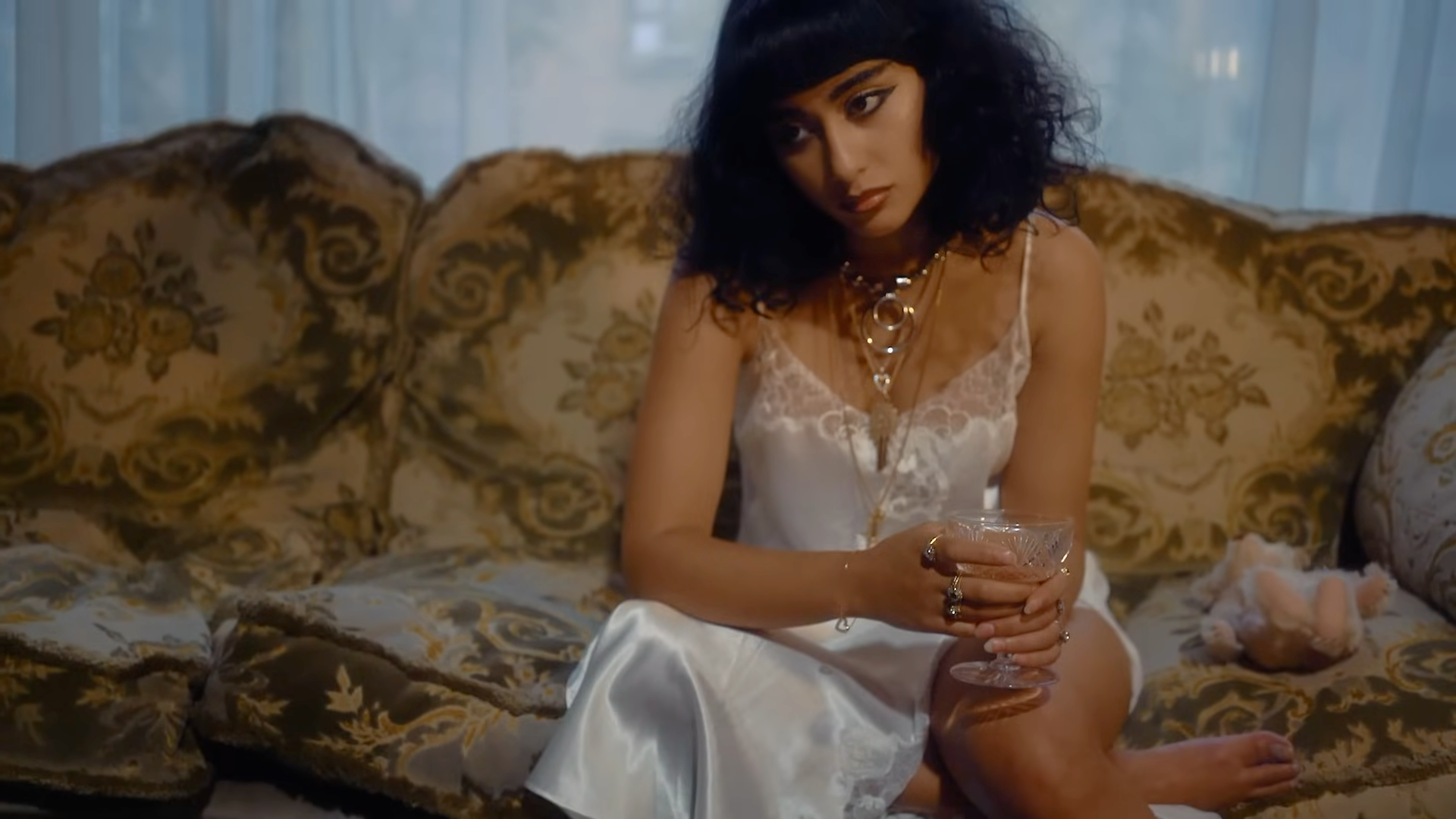 Woman in a white dress sitting on a patterned couch holding a glass.